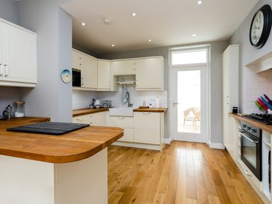 A kitchen with wooden countertops white cabinets a stove a sink a door leading outside and a clock on the wall at The Beach House Alnmouth