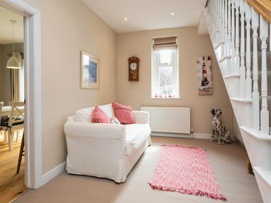 A living area with a white sofa with red striped pillows a wall clock window radiator a pink rug and a staircase with a dog figurine at The Beach House in Alnmouth