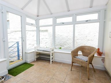 A conservatory with tiled floor a wicker chair with a cushion a white bench with a cushion and a glass door at The Beach House Alnmouth