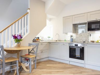 A kitchen with light gray cabinets wooden floor a small wooden table with two chairs and flowers in a vase under a staircase at Wisteria Cottage in Warkworth