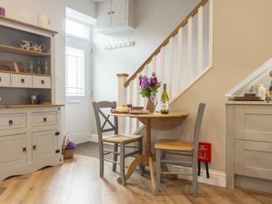 A small dining area with a round wooden table and two chairs near a white door and a staircase with a wooden handrail at Wisteria Cottage in Warkworth