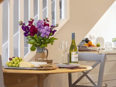 A dining table with a vase of purple flowers grapes cheese crackers a wine bottle and an empty wine glass at Wisteria Cottage in Warkworth