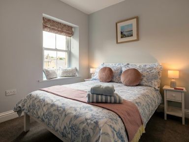 A bedroom with a bed with floral bedding and round pillows bedside tables with lamps and a window with cushions at Wisteria Cottage in Warkworth