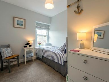 A bedroom with a single bed striped bedding a chair bedside table with a lamp and a dresser with a lamp and mirror at Wisteria Cottage in Warkworth