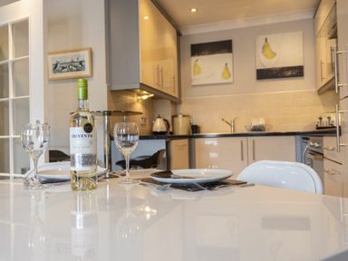 A dining table with a bottle of wine and glasses in a kitchen with cabinets and wall art at Puffin View in Seahouses