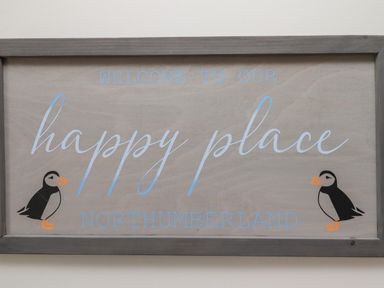 A wooden sign with the text welcome to our happy place northumberland and two puffin illustrations
