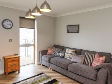 A living room with a gray sofa with multiple cushions a wooden side table and a wall clock at Puffin View in Seahouses