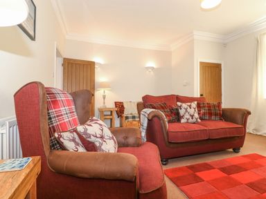 A living room with red upholstered armchair and sofa with patterned cushions a red checkered rug and wooden side tables at Willow Cottage in Lesbury