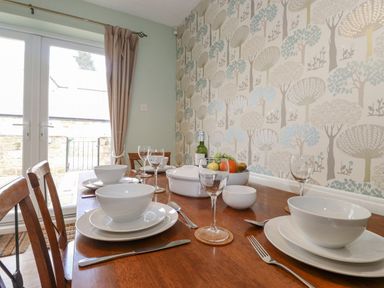 A dining room with a wooden table set for four people with white dishes glasses and cutlery next to a wall with tree patterned wallpaper at Willow Cottage in Lesbury