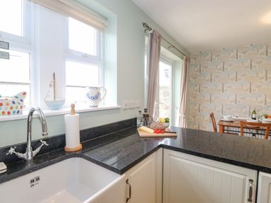 A kitchen sink with faucet and paper towel holder on a black countertop beside a window and a dining table with chairs in the background at Willow Cottage in Lesbury