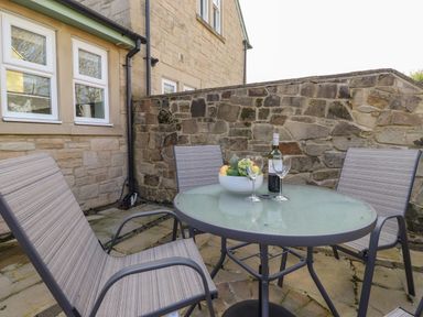 An outdoor patio area with a glass table and three chairs a bowl of fruit and a bottle of wine on the table at Willow Cottage in Lesbury