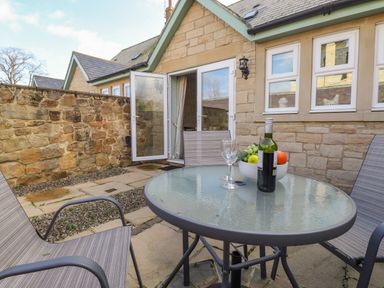 An outdoor patio with a glass table and chairs a bottle of wine and a bowl of fruit at Willow Cottage in Lesbury