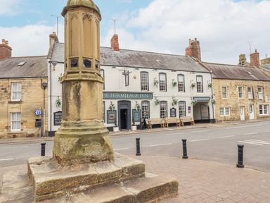 A street view with a stone monument in the foreground and The Hermitage Inn building with benches and flower baskets in Warkworth