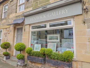 The exterior of Fenwick Gallery with stone walls a window displaying framed artwork and green topiary plants in pots Warkworth