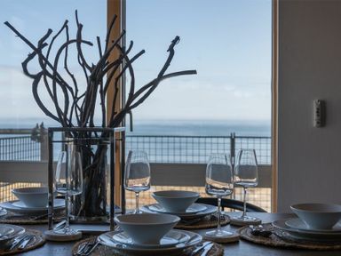 A dining table set with bowls plates wine glasses and a centerpiece of branches by a window with ocean view at Keildon in Seahouses