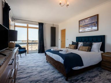 A bedroom with a large bed, blue chair, dresser with lamp, and a window with a view at Keildon in Seahouses