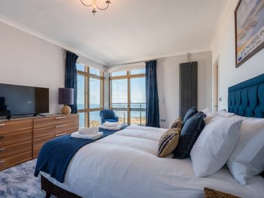 A bedroom with a bed pillows folded towels a wooden dresser a lamp a blue chair and large windows overlooking water at Keildon in Seahouses