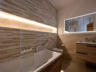 A bathroom with a bathtub a wall-mounted toilet a backlit mirror and a wooden vanity at Keildon in Seahouses