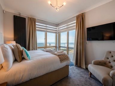 A bedroom with a bed beside windows showing a sea view and a chair near a wall mounted TV at Keildon in Seahouses