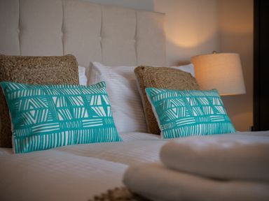 A bed with white linens two woven pillows turquoise patterned cushions and a bedside lamp at Keildon in Seahouses