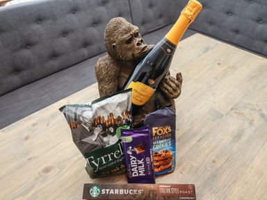 A bottle of prosecco held by a gorilla statue with packets of Tyrrells crisps, Cadbury Dairy Milk chocolate, Fox's milk chocolate cookies and Starbucks Italian Style Roast coffee on a wooden table
