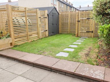 A small garden with grass, stepping stones, wooden fence, a gate, and a black storage shed at Muscle Shoals in Seahouses