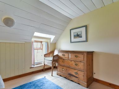 A small room with a wooden chest of drawers a wicker chair a window with curtains and a blue rug on the floor at Sandpiper Cottage Low Newton-by-the-Sea