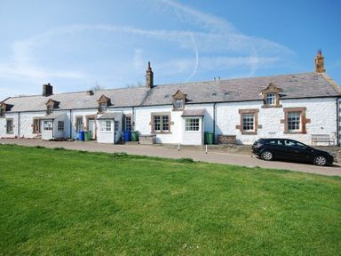 A row of white cottages with small porches and chimneys next to a driveway with a black car and green lawn at Sandpiper Cottage Low Newton-by-the-Sea
