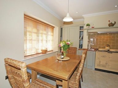 A dining area with a wooden table and wicker chairs next to a window with blinds and a kitchen with beige cabinets and an Aga cooker at Grange Cottage in Alnmouth