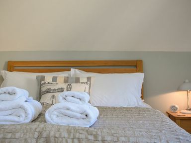 A bed with white pillows and folded towels two cushions and a nightstand with a lamp and clock at Grange Cottage in Alnmouth