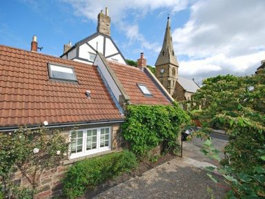 A cottage with red roof tiles and skylights next to a church with a clock tower and trees at Grange Cottage in Alnmouth