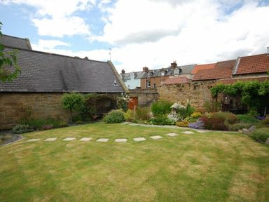 A garden with a stone path lawn stone walls plants and houses in the background at Grange Cottage in Alnmouth