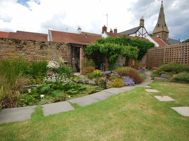 A garden with a stone pathway a pond plants and flowers a stone wall a wooden trellis and a building with a church steeple in the background at Grange Cottage in Alnmouth