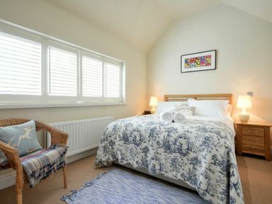 A bedroom with a bed with patterned bedding two lamps on wooden bedside tables a wicker chair with cushions and a window with blinds at Martins Cottage in Alnmouth
