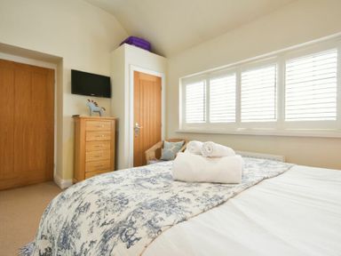 A bedroom with a bed covered in a patterned blanket and rolled towels windows with shutters a wooden chest of drawers a wall-mounted television a wicker chair and two wooden doors at Martins Cottage in Alnmouth