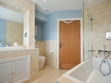 A bathroom with a bathtub with glass screen a sink with cabinets a toilet and a wooden door at Martins Cottage in Alnmouth