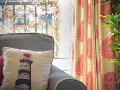 A cushion with a lighthouse design on a grey armchair next to floral patterned curtains and a plant at The Boathouse (Alnmouth) in Alnmouth