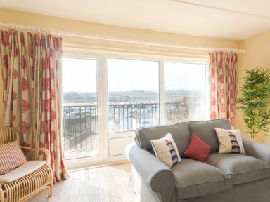 A living room with a gray sofa and patterned curtains by large windows overlooking a marina at The Boathouse (Alnmouth) in Alnmouth