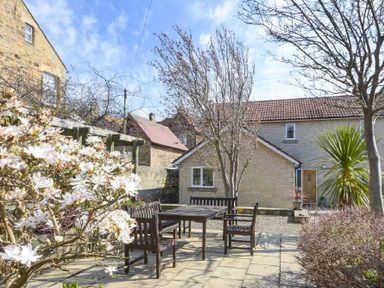 An outdoor patio area with a wooden table and chairs surrounded by flowering bushes and trees at The Boathouse in Alnmouth