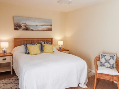 A bedroom with a wooden bed frame white bedding and cushions two bedside tables with lamps and a wicker chair at Coach House (Alnmouth) in Alnmouth