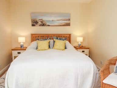 A bedroom with a double bed with yellow and blue pillows two bedside tables with lamps and a wicker chair at Coach House (Alnmouth) in Alnmouth
