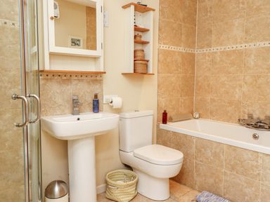 A bathroom with a pedestal sink, toilet, bathtub, wall shelves with baskets, mirror cabinet, and a glass shower door at Coach House (Alnmouth) in Alnmouth