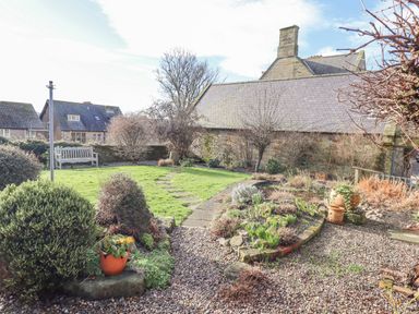 A garden with bushes, a path, flower beds, a bench, and stone buildings in the background at Coach House (Alnmouth) in Alnmouth