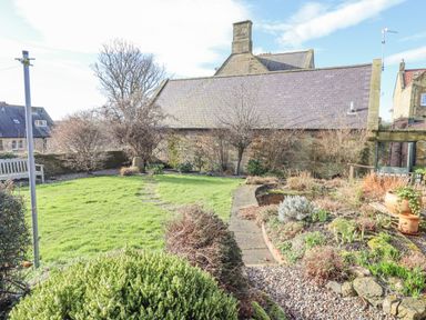 A garden with grass, shrubs, a stone path, and a stone building with slate roof in the background at Coach House (Alnmouth) in Alnmouth