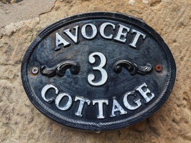 A black oval house number plaque with white letters and the number 3 on a stone wall at Avocet Cottage Bamburgh