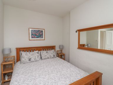 A bedroom with a wooden bed frame, patterned bedspread, bedside tables with lamps, a wall mirror, and a floral picture at Avocet Cottage in Bamburgh