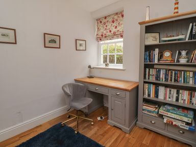A study room with a gray desk and chair next to a window and a bookcase filled with books and puzzles at Avocet Cottage Bamburgh