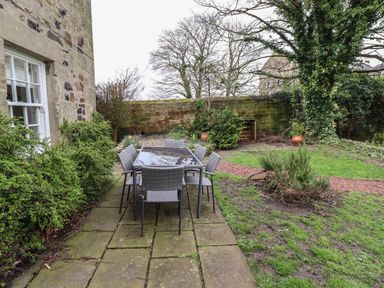 An outdoor patio with a glass table and six chairs beside a stone house and a garden with trees at Avocet Cottage in Bamburgh