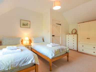 A bedroom with two single beds a nightstand with a lamp a chest of drawers a wardrobe and a door at Dunlin Cottage in Bamburgh