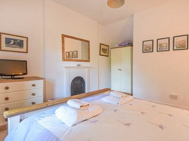 A bedroom with a double bed and towels on it a white dresser with a television a white wardrobe a fireplace with a mirror above it and framed pictures on the walls at Dunlin Cottage in Bamburgh